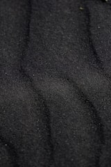 Dark, fine-textured sand displays soft, undulating patterns across its surface. The sand has a deep, almost black hue and subtle shadow lines create a wavy appearance.