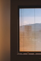 A window covered with horizontal blinds partially open to reveal an exterior view. The scenery outside is slightly blurred, featuring shades of the sky transitioning from blue at the top to warmer colors near the bottom, possibly indicating sunset or sunrise. The interior wall surrounding the window is dimly lit.