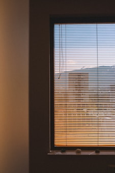 A window covered with horizontal blinds partially open to reveal an exterior view. The scenery outside is slightly blurred, featuring shades of the sky transitioning from blue at the top to warmer colors near the bottom, possibly indicating sunset or sunrise. The interior wall surrounding the window is dimly lit.