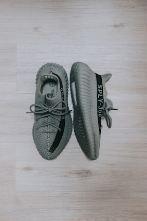 A pair of gray athletic shoes with a distinctive design. The shoes feature a textured pattern and a stripe with text along the side. The sneakers are placed on a light wooden floor, with one shoe lying flat and the other tilted to show the sole.