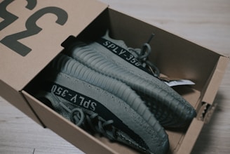 A pair of gray sneakers with the label 'SPLY-350' is placed inside a cardboard shoe box on a light wooden floor. The sneakers have a ribbed design on the sole and are partially visible due to the lid of the box being slightly open.