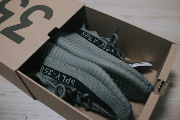 A pair of gray sneakers with the label 'SPLY-350' is placed inside a cardboard shoe box on a light wooden floor. The sneakers have a ribbed design on the sole and are partially visible due to the lid of the box being slightly open.