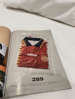 A magazine page features a red soccer jersey with yellow and dark blue accents, encased in a transparent plastic bag. The jersey is branded with 'adidas' and has a badge resembling the Spanish national team's emblem on the chest. The page is numbered 289 and includes some text references and credits at the bottom.