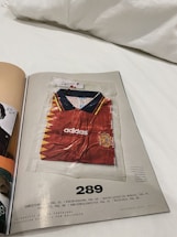 A magazine page features a red soccer jersey with yellow and dark blue accents, encased in a transparent plastic bag. The jersey is branded with 'adidas' and has a badge resembling the Spanish national team's emblem on the chest. The page is numbered 289 and includes some text references and credits at the bottom.
