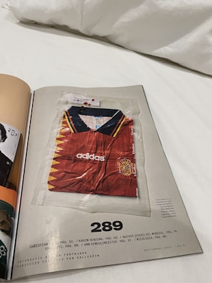 A magazine page features a red soccer jersey with yellow and dark blue accents, encased in a transparent plastic bag. The jersey is branded with 'adidas' and has a badge resembling the Spanish national team's emblem on the chest. The page is numbered 289 and includes some text references and credits at the bottom.