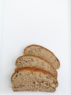Three slices of brown bread are arranged on a white surface. The bread has a dense and slightly grainy texture with visible seeds and a golden brown crust.