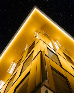 A modern building with sleek, angular architecture features prominent yellow lighting that contrasts against a dark night sky dotted with stars. The building's façade is composed of geometric panels, giving it a futuristic appearance.