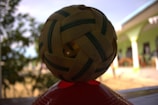 A handwoven ball with a checkered pattern sits on top of a red base. The ball features alternating strips of light brown and dark green. In the background, there are blurred trees and part of a building with green columns and white accents.