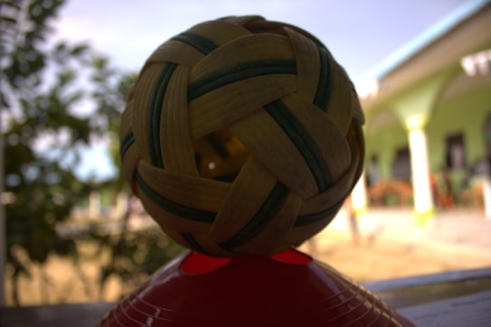 A handwoven ball with a checkered pattern sits on top of a red base. The ball features alternating strips of light brown and dark green. In the background, there are blurred trees and part of a building with green columns and white accents.