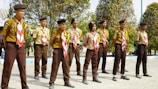 A group of children dressed in scout uniforms stands outdoors in a line, wearing brown berets and distinctive red and white neckerchiefs. They appear focused and attentive, possibly participating in a scouting event or ceremony. The children are positioned on a paved area, with trees and a few buildings in the background.