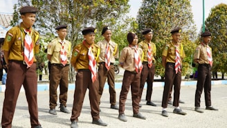 A group of children dressed in scout uniforms stands outdoors in a line, wearing brown berets and distinctive red and white neckerchiefs. They appear focused and attentive, possibly participating in a scouting event or ceremony. The children are positioned on a paved area, with trees and a few buildings in the background.