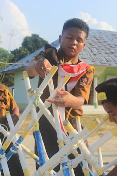 A person in a scout uniform is assembling a structure made from interconnected sticks, which are painted in yellow, white, and blue. Two other individuals, also in scout uniforms, are partially visible in the scene. The setting appears to be outdoors near a building with a rusty metal roof. Trees are visible in the background under a blue sky with a few clouds.