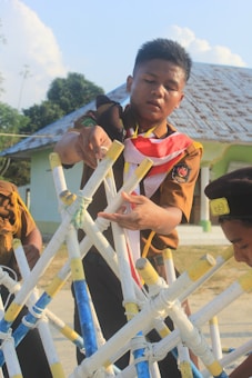 A person in a scout uniform is assembling a structure made from interconnected sticks, which are painted in yellow, white, and blue. Two other individuals, also in scout uniforms, are partially visible in the scene. The setting appears to be outdoors near a building with a rusty metal roof. Trees are visible in the background under a blue sky with a few clouds.