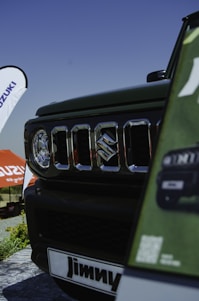 A close-up view of a vehicle's front grille featuring the Suzuki logo. The background shows a banner and an umbrella with the Suzuki branding. The view includes part of the car and signage displaying the name 'Jimny.' The setting suggests an outdoor event or exhibition under a clear blue sky.