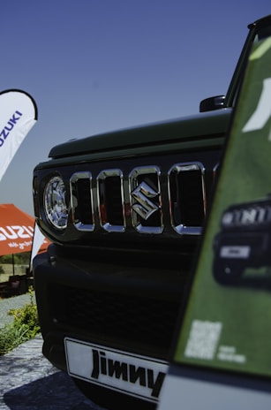 A close-up view of a vehicle's front grille featuring the Suzuki logo. The background shows a banner and an umbrella with the Suzuki branding. The view includes part of the car and signage displaying the name 'Jimny.' The setting suggests an outdoor event or exhibition under a clear blue sky.