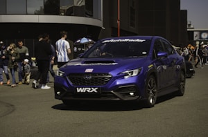 A vibrant blue sports sedan with the brand 'WRX' on the front is parked on a pavement. It has advertising decals, including 'AutoTrader' on the front windshield. People are gathered around nearby, some walking, others standing and observing. Some are dressed casually, with one person holding an umbrella featuring an automobile brand logo. The backdrop features a modern building with large windows.
