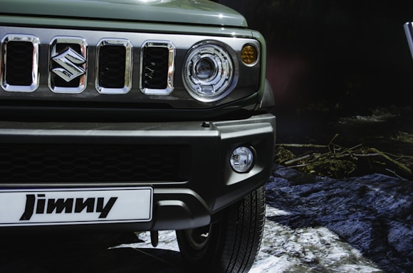 Close-up of a vehicle's front portion featuring a detailed view of the grille, headlights, and bumper. The logo, a prominent S, is clearly visible in the center of the grille. The setting appears to be outdoors with rocks and vegetation faintly visible in the background.
