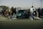 A gathering of people surrounds a green sports car parked in an open area. The scene includes several individuals walking and interacting around the vehicle, with some pushing strollers. A fence stretches along the background beneath a clear sky, suggesting an outdoor setting.
