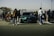 A gathering of people surrounds a green sports car parked in an open area. The scene includes several individuals walking and interacting around the vehicle, with some pushing strollers. A fence stretches along the background beneath a clear sky, suggesting an outdoor setting.