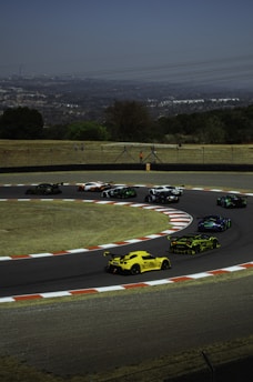 Several sports cars are seen racing on a track with a sharp turn. The background features a green grassy area and a few trees, while a hazy cityscape can be seen in the distance under a clear sky.