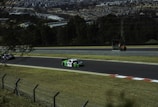 Cars racing on a track with a view of a town in the background. The scene includes two cars on a delineated raceway, bordered by grass and a fence. In the background, there is a group of people in an official-looking area alongside the track, and trees leading to a distant view of urban buildings.