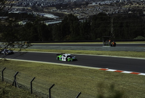 Cars racing on a track with a view of a town in the background. The scene includes two cars on a delineated raceway, bordered by grass and a fence. In the background, there is a group of people in an official-looking area alongside the track, and trees leading to a distant view of urban buildings.