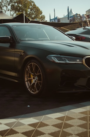 A sleek, dark green luxury sports car is parked on a checkered floor. The car has a shiny exterior with detailed gold and brown rims. In the background, there are tents and flags, suggesting an outdoor event or car show. Trees can be seen in the distance, adding a natural element to the setting.