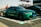 A vibrant green sports sedan is prominently displayed in an outdoor setting. The car features sleek, aerodynamic lines and a shiny finish, parked on a checkered mat. People are visible in the background, standing near other vehicles with banners and a building structure in view.