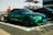 A vibrant green sports sedan is prominently displayed in an outdoor setting. The car features sleek, aerodynamic lines and a shiny finish, parked on a checkered mat. People are visible in the background, standing near other vehicles with banners and a building structure in view.