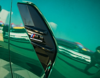 Close-up of a shiny green car surface with a BMW M Competition emblem. The reflection on the car's surface shows blurred colors and silhouettes, suggesting an outdoor setting possibly near a racetrack or event.