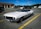 A classic white convertible car is parked in an empty parking lot. Its top is down, showcasing its vintage design with a sleek, elongated hood and round headlights. Several other modern cars are parked in the background near a building with a service area.