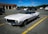A classic white convertible car is parked in an empty parking lot. Its top is down, showcasing its vintage design with a sleek, elongated hood and round headlights. Several other modern cars are parked in the background near a building with a service area.