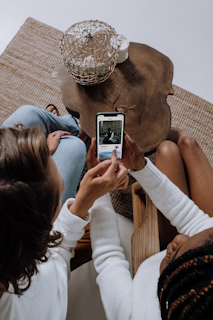 Two people are looking at a smartphone together, sitting on chairs next to a wooden coffee table. A decorative woven basket is placed on the table, along with a small item inside. They appear to be engaged with the content on the phone, possibly discussing or sharing something interesting.