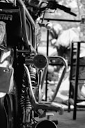Close-up view of a motorcycle parked in a garage or workshop, with shiny chrome parts and detailed mechanical components in focus. In the background, there are blurred figures working or interacting with another motorcycle.