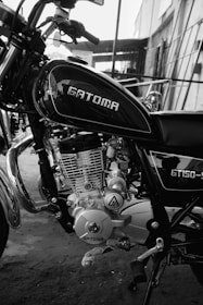 A black and white photo of a motorcycle, prominently displaying the engine and body. The motorcycle is branded 'GATOMA' with the model 'GT150' visible. It appears to be in a workshop or garage setting, with some building structures in the background.