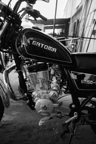 A black and white photo of a motorcycle, prominently displaying the engine and body. The motorcycle is branded 'GATOMA' with the model 'GT150' visible. It appears to be in a workshop or garage setting, with some building structures in the background.
