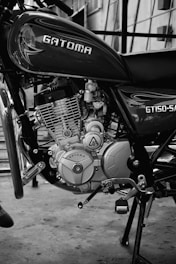 A black and white photograph focusing on the engine and lower body of a Gatoma GT150-SA motorcycle. The engine components, including the intricate parts and the Gatoma logo, are clearly visible. The background shows a concrete floor and a building with stairway railings.