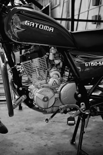 A black and white photograph focusing on the engine and lower body of a Gatoma GT150-SA motorcycle. The engine components, including the intricate parts and the Gatoma logo, are clearly visible. The background shows a concrete floor and a building with stairway railings.