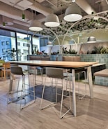 A modern office lounge area features a long wooden table with metal legs surrounded by high chairs. Overhead, white dome lights hang from an industrial-style ceiling. The space is decorated with green plants and partitioned by large glass windows, allowing natural light to enter. The floor is covered with light gray wood-like tiles, and cushioned benches with green-tiled backing provide additional seating.
