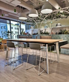 A modern office lounge area features a long wooden table with metal legs surrounded by high chairs. Overhead, white dome lights hang from an industrial-style ceiling. The space is decorated with green plants and partitioned by large glass windows, allowing natural light to enter. The floor is covered with light gray wood-like tiles, and cushioned benches with green-tiled backing provide additional seating.