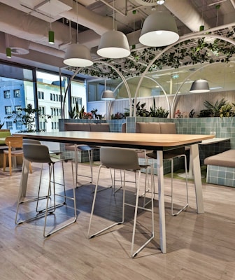 A modern office lounge area features a long wooden table with metal legs surrounded by high chairs. Overhead, white dome lights hang from an industrial-style ceiling. The space is decorated with green plants and partitioned by large glass windows, allowing natural light to enter. The floor is covered with light gray wood-like tiles, and cushioned benches with green-tiled backing provide additional seating.
