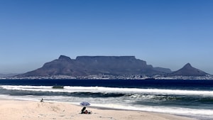 A scenic beach view with waves gently rolling onto the sand. In the distance, a prominent flat-topped mountain dominates the skyline, with a cityscape spread across the base of the mountain. A beachgoer sits under a blue umbrella, while another person can be seen walking along the beach.