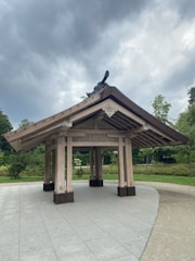 A wooden pavilion structure with a traditional architectural design, featuring a gabled roof and intricate details on the eaves. It stands on a paved surface surrounded by greenery, including trees and bushes. The sky above is overcast with thick, grey clouds.