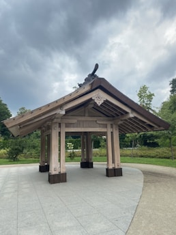 A wooden pavilion structure with a traditional architectural design, featuring a gabled roof and intricate details on the eaves. It stands on a paved surface surrounded by greenery, including trees and bushes. The sky above is overcast with thick, grey clouds.