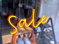 Bright neon signage spells out the word 'Sale', suspended in front of a shop window. The interior shows pairs of shoes displayed on an orange, tiered rack.
