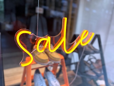 Bright neon signage spells out the word 'Sale', suspended in front of a shop window. The interior shows pairs of shoes displayed on an orange, tiered rack.