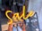 Bright neon signage spells out the word 'Sale', suspended in front of a shop window. The interior shows pairs of shoes displayed on an orange, tiered rack.