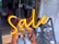 Bright neon signage spells out the word 'Sale', suspended in front of a shop window. The interior shows pairs of shoes displayed on an orange, tiered rack.