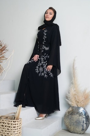 A woman dressed in an elegant black gown with intricate white floral embroidery stands gracefully on a set of stairs. Her hair is partially covered with a black headscarf, and she is wearing metallic silver heels. Nearby, there are decorative elements, including a textured large vase and dried plants, contributing to a sophisticated and stylish ambiance.