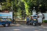A man stands near a blue and red tuk-tuk parked in front of an ornate, closed iron gate. The area is surrounded by lush green trees and there is another tuk-tuk parked nearby. The architecture has a historic feel, and sunlight filters through the trees, casting dappled shadows on the ground.